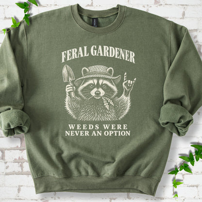 Military Green classic sweatshirt laid flat, showing a cream raccoon in a straw hat holding a trowel, with "Feral Gardener" and "Weeds Were Never an Option" lettering.