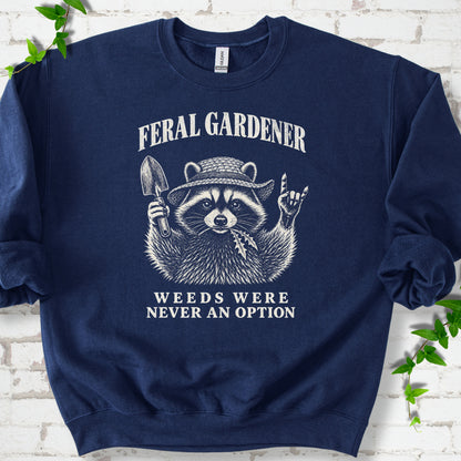 Navy classic sweatshirt laid flat, showing a cream raccoon in a straw hat holding a trowel, with "Feral Gardener" and "Weeds Were Never an Option" lettering.