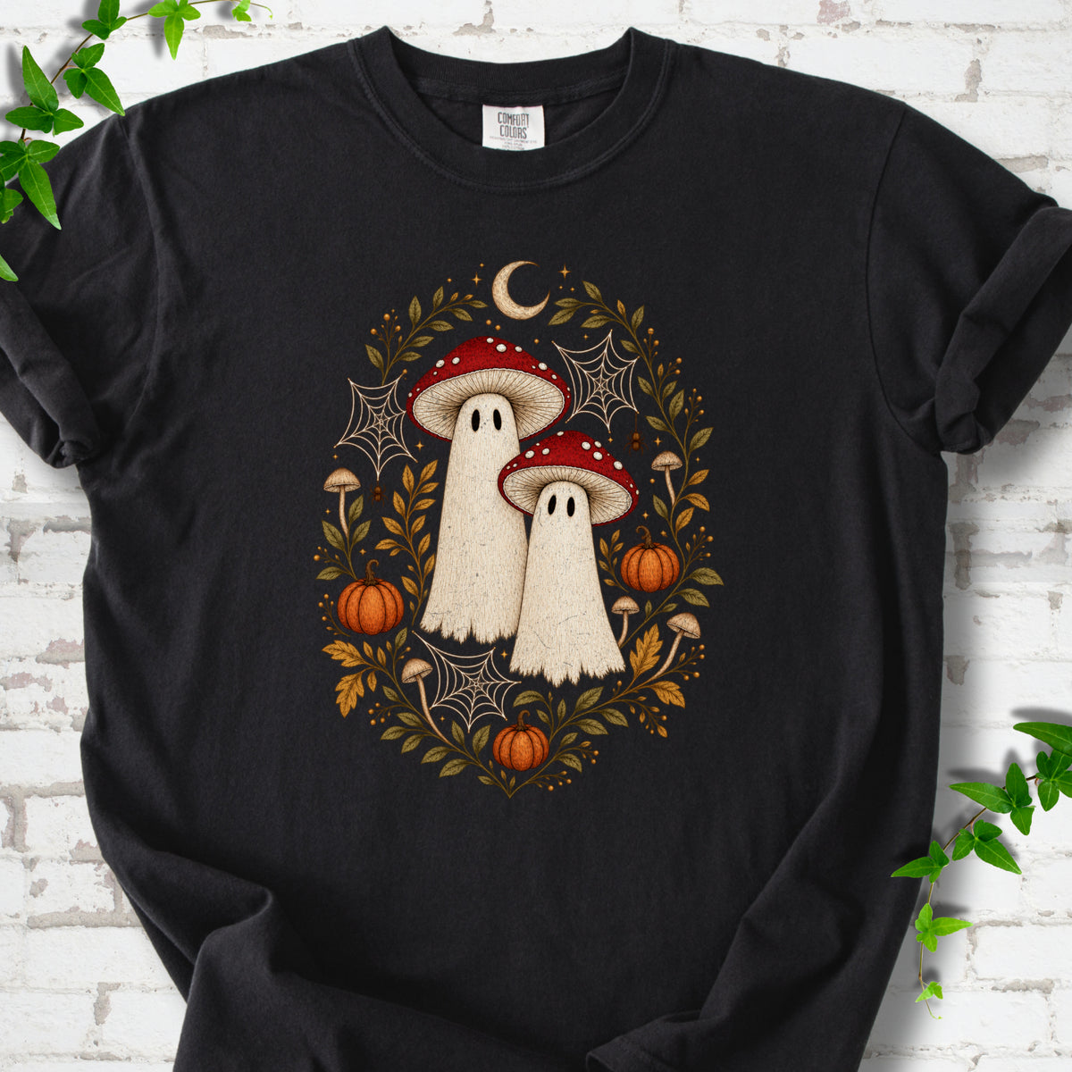 Black Comfort Colors® T-shirt laid flat, showing two cream ghosts wearing red mushroom caps, surrounded by small pumpkins, autumn leaves and a crescent moon.