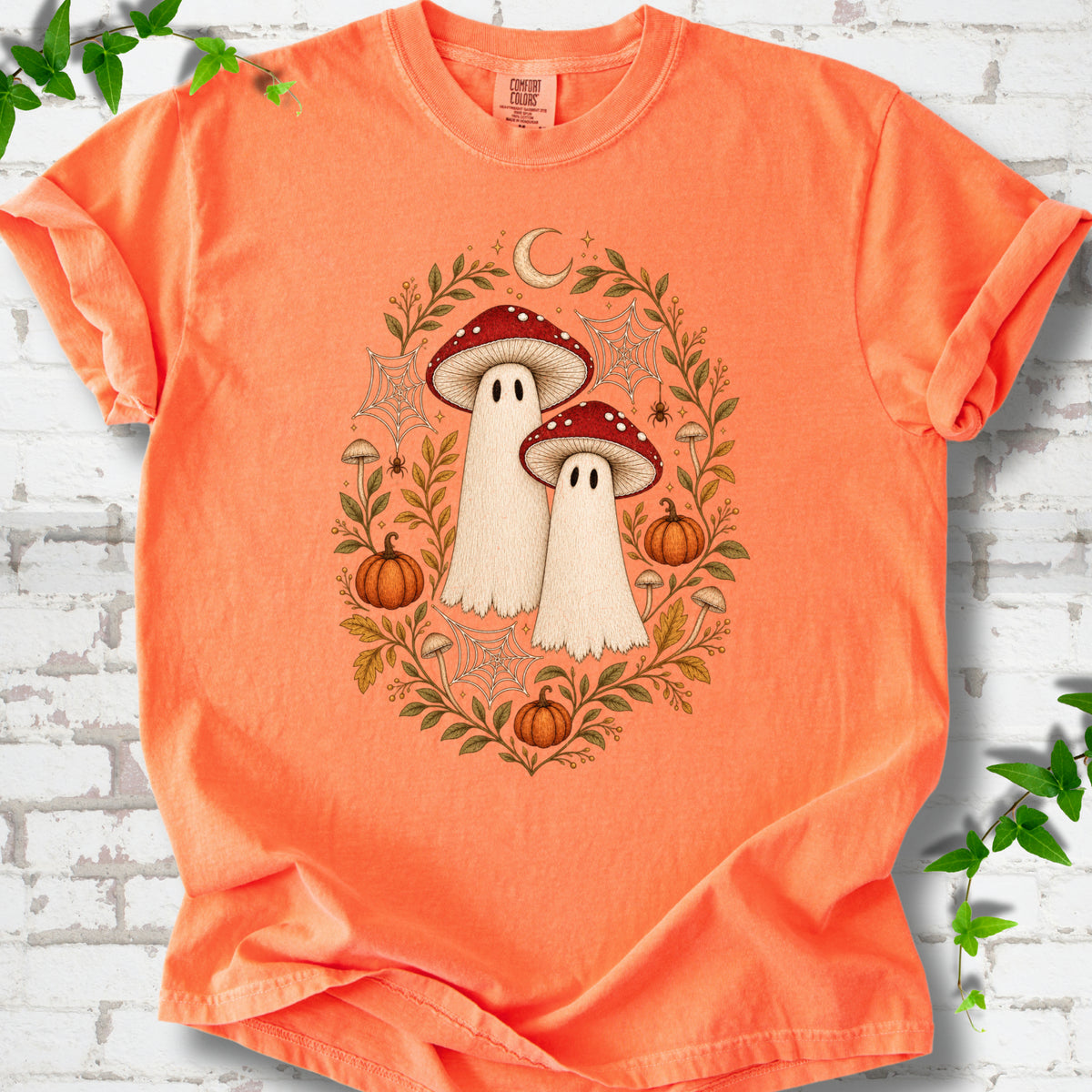 Melon Comfort Colors® T-shirt laid flat, showing two cream ghosts wearing red mushroom caps, surrounded by small pumpkins, autumn leaves and a crescent moon.
