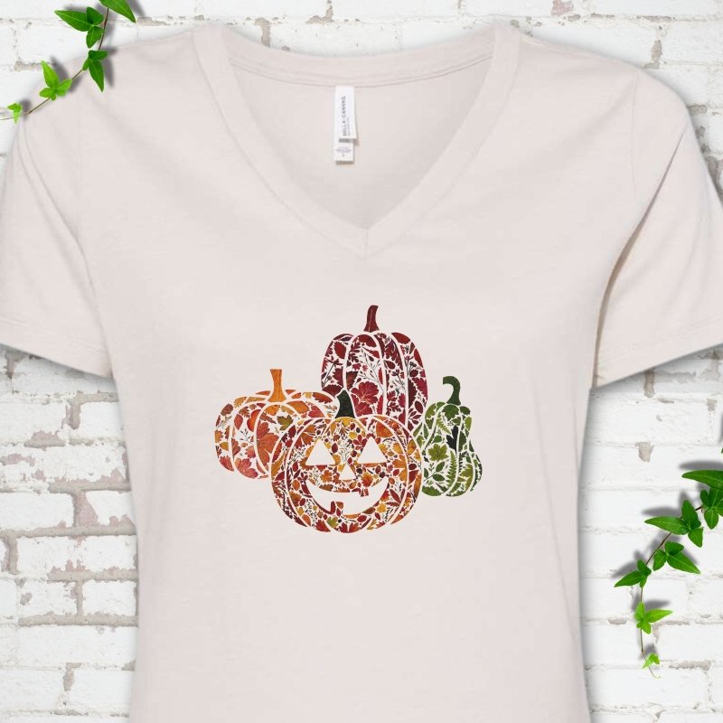 Heather Dust Bella Canvas® ladies' V-neck T-shirt flat lay featuring four pumpkins filled with colorful autumn leaves, ferns and wildflowers, including one smiling jack-o'-lantern.