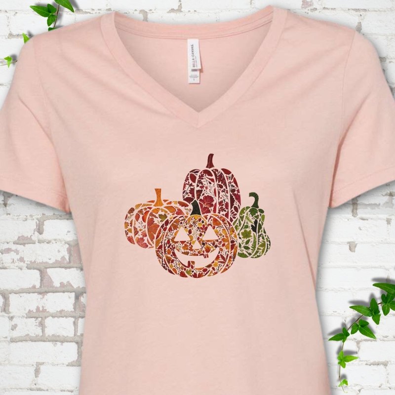 Heather Peach Bella Canvas® ladies' V-neck T-shirt flat lay featuring four pumpkins filled with colorful autumn leaves, ferns and wildflowers, including one smiling jack-o'-lantern.