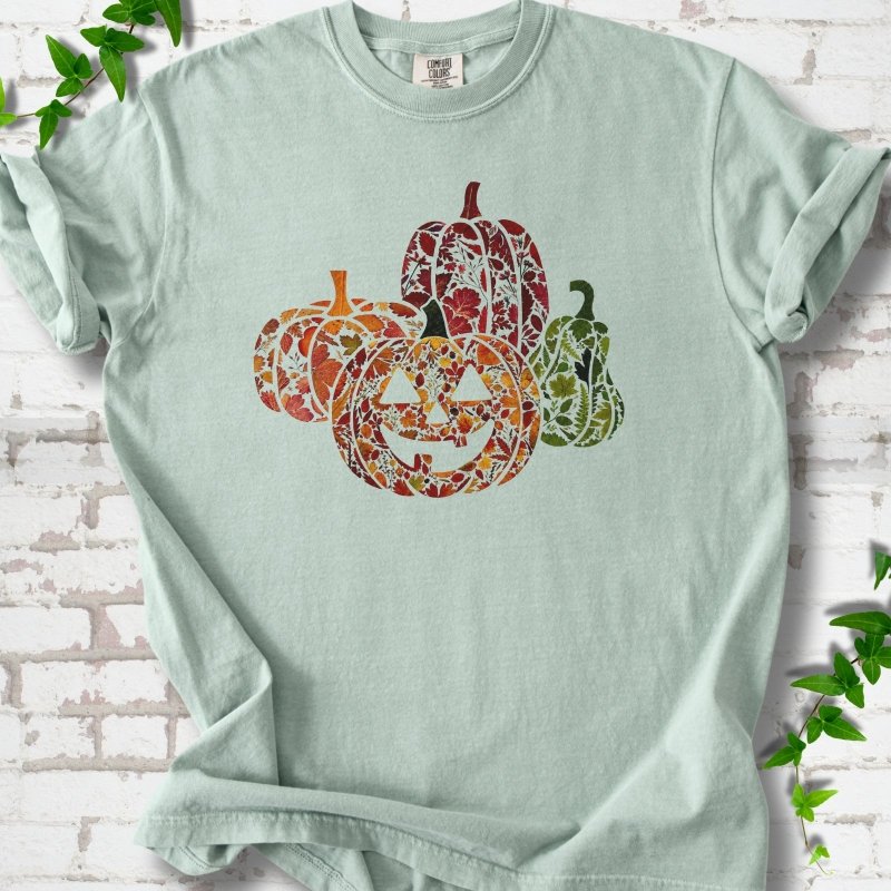 Bay Comfort Colors® T-shirt flat lay featuring four pumpkins filled with colorful autumn leaves, ferns and wildflowers, including one smiling jack-o'-lantern.