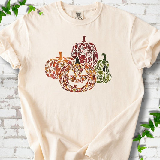 Ivory Comfort Colors® T-shirt flat lay featuring four pumpkins filled with colorful autumn leaves, ferns and wildflowers, including one smiling jack-o'-lantern.