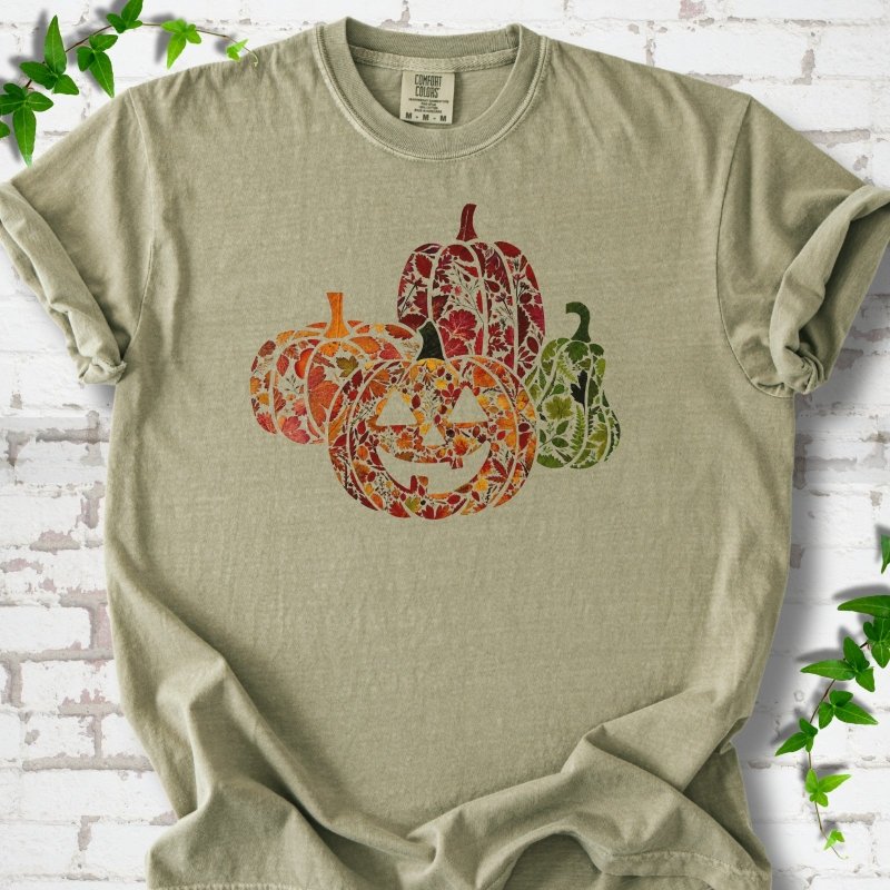 Khaki Comfort Colors® T-shirt flat lay featuring four pumpkins filled with colorful autumn leaves, ferns and wildflowers, including one smiling jack-o'-lantern.