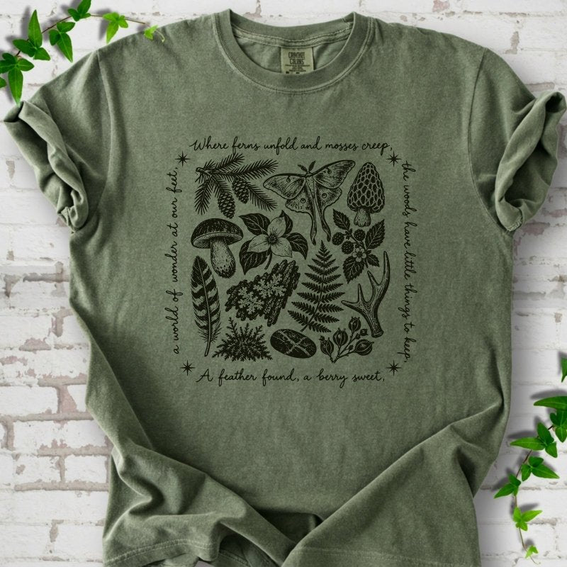 Moss green short-sleeve tee with woodland finds engraving in dark green ink, framed by "a world of wonder at our feet".