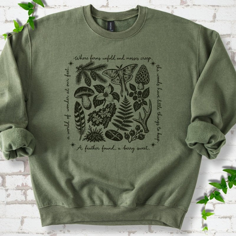 Military green crewneck sweatshirt with woodland finds engraving in dark ink, framed by "a world of wonder at our feet".