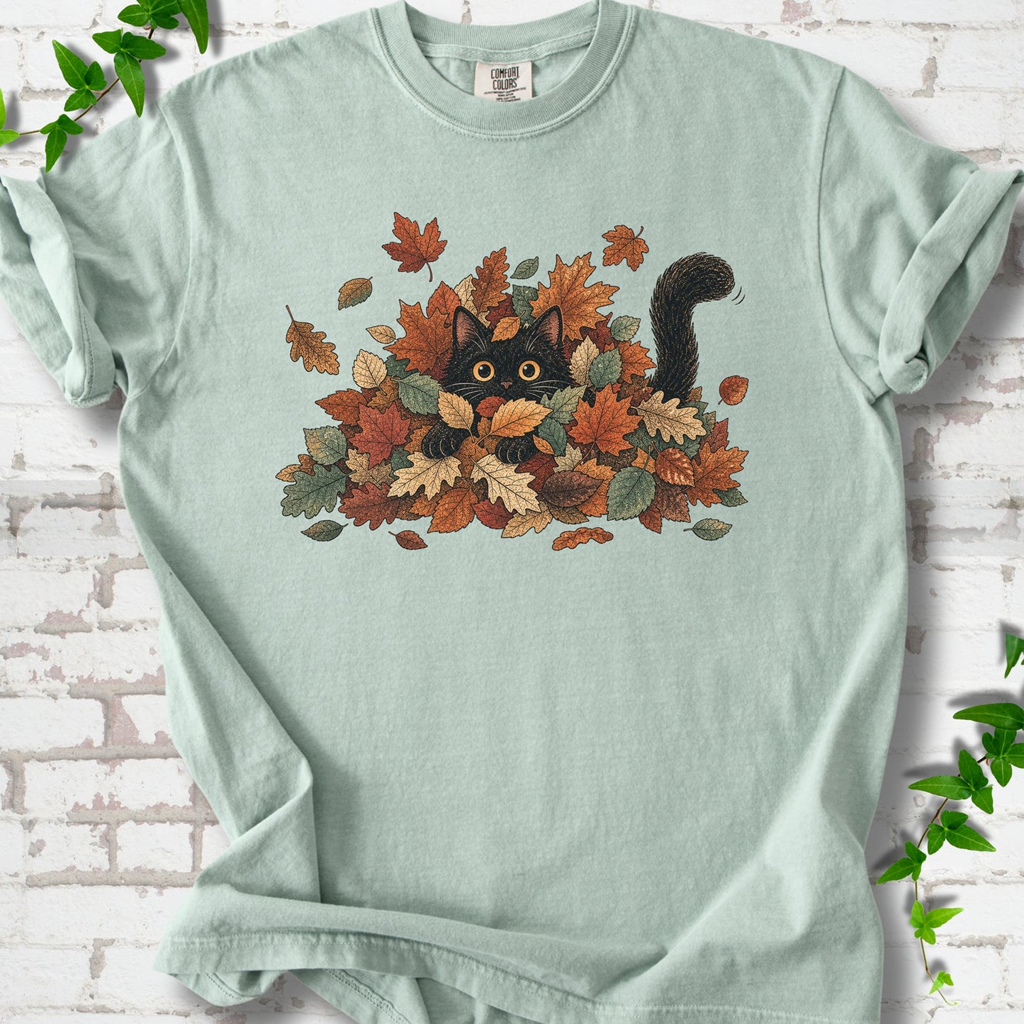 Bay Comfort Colors® T-shirt flat lay featuring a black cat peeking from a pile of orange and green autumn leaves.
