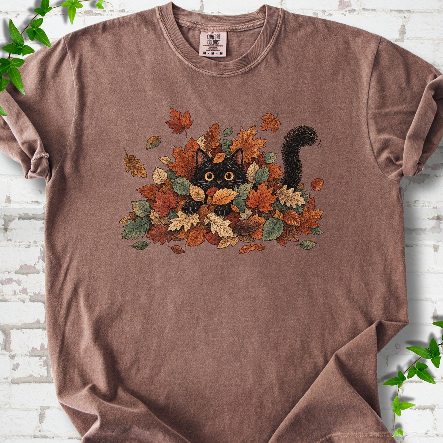 Espresso Comfort Colors® T-shirt flat lay featuring a black cat peeking from a pile of orange and green autumn leaves.