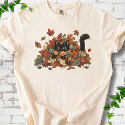 Ivory Comfort Colors® T-shirt flat lay featuring a black cat peeking from a pile of orange and green autumn leaves.