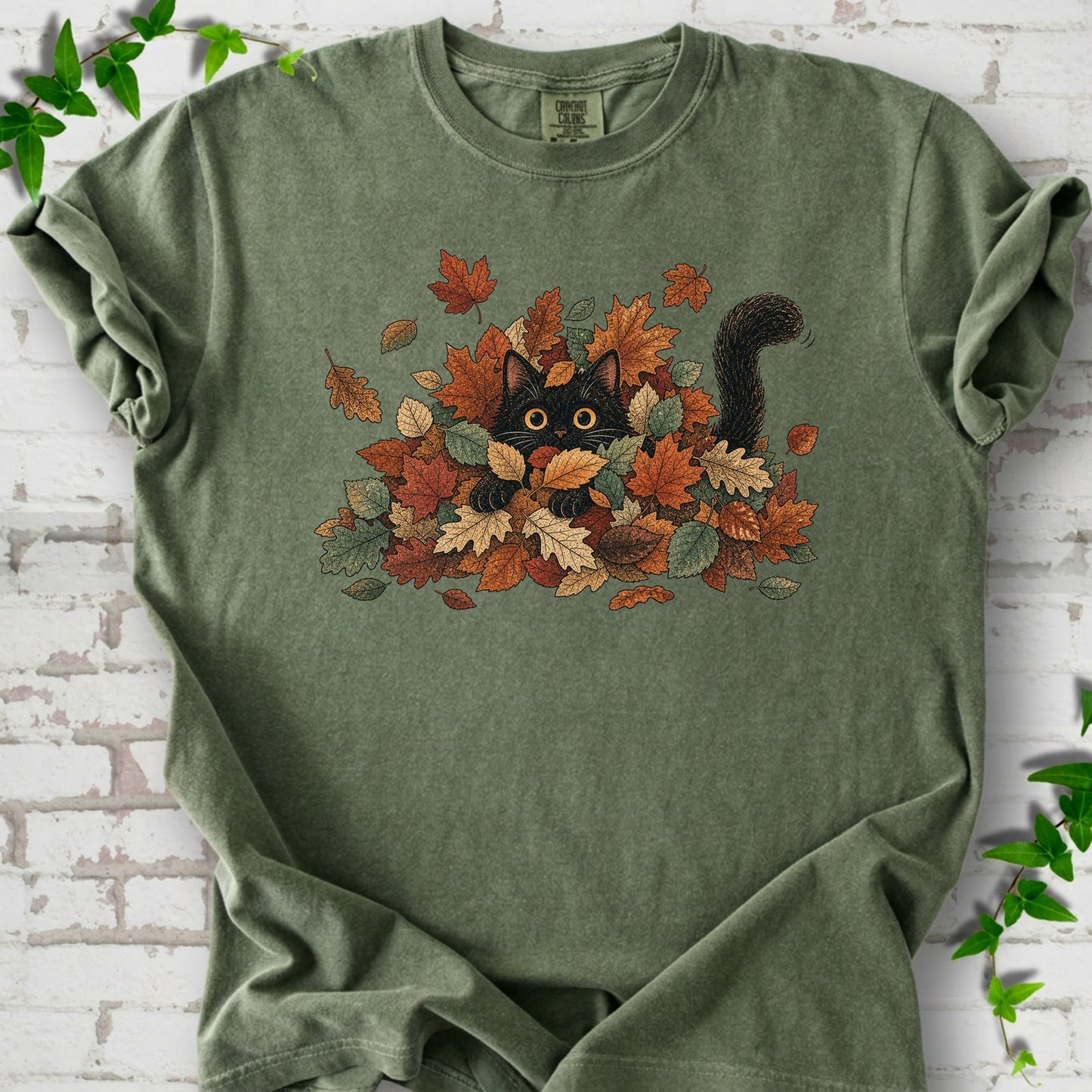 Moss Comfort Colors® T-shirt flat lay featuring a black cat peeking from a pile of orange and green autumn leaves.