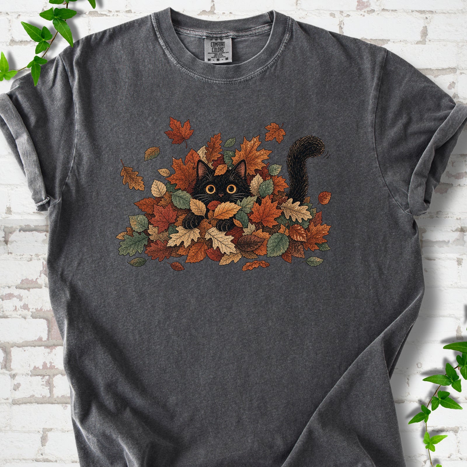 Pepper Comfort Colors® T-shirt flat lay featuring a black cat peeking from a pile of orange and green autumn leaves.