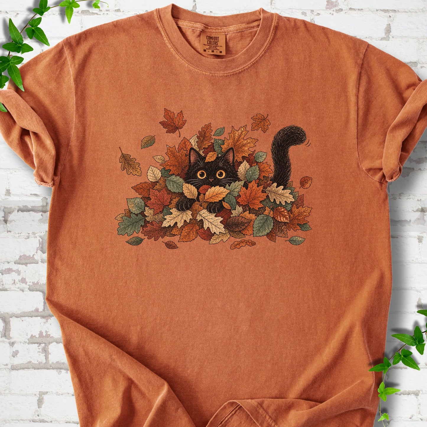 Yam Comfort Colors® T-shirt flat lay featuring a black cat peeking from a pile of orange and green autumn leaves.