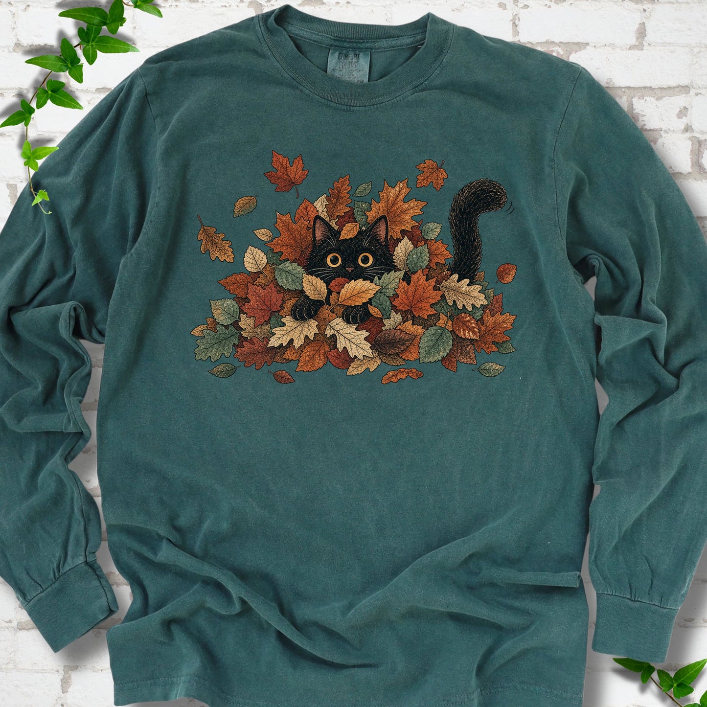 Blue Spruce Comfort Colors® long sleeve T-shirt flat lay featuring a black cat peeking from a pile of orange and green autumn leaves.