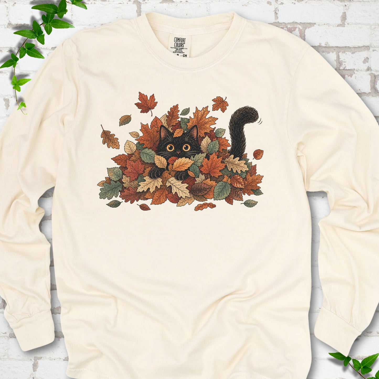 Ivory Comfort Colors® long sleeve T-shirt flat lay featuring a black cat peeking from a pile of orange and green autumn leaves.