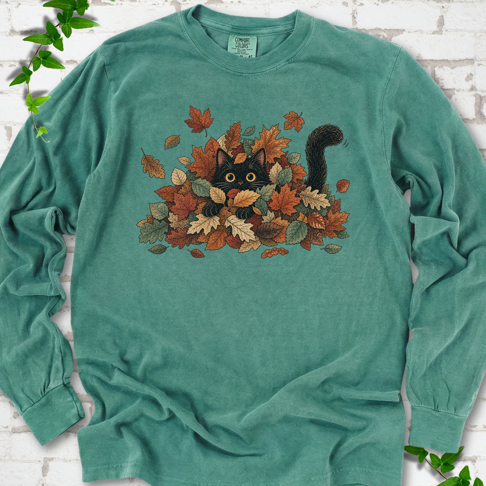 Light Green Comfort Colors® long sleeve T-shirt flat lay featuring a black cat peeking from a pile of orange and green autumn leaves.