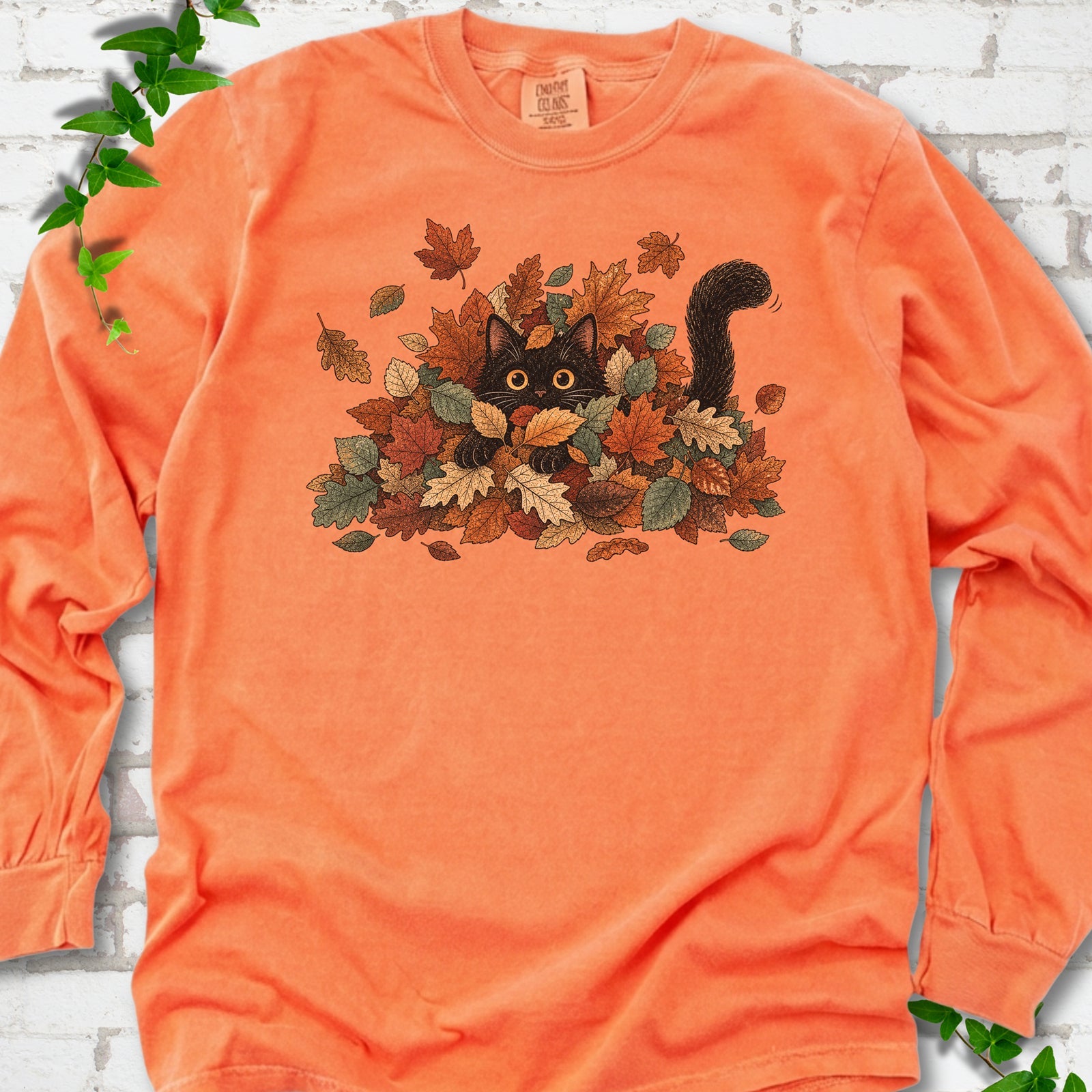 Melon Comfort Colors® long sleeve T-shirt flat lay featuring a black cat peeking from a pile of orange and green autumn leaves.