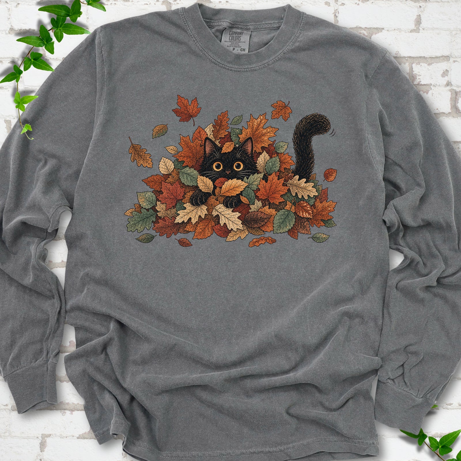 Pepper Comfort Colors® long sleeve T-shirt flat lay featuring a black cat peeking from a pile of orange and green autumn leaves.