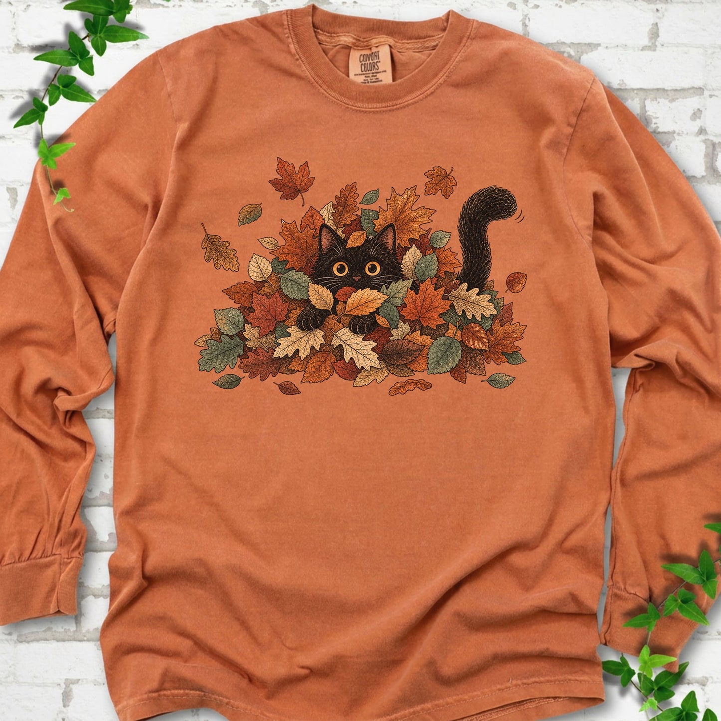 Yam Comfort Colors® long sleeve T-shirt flat lay featuring a black cat peeking from a pile of orange and green autumn leaves.