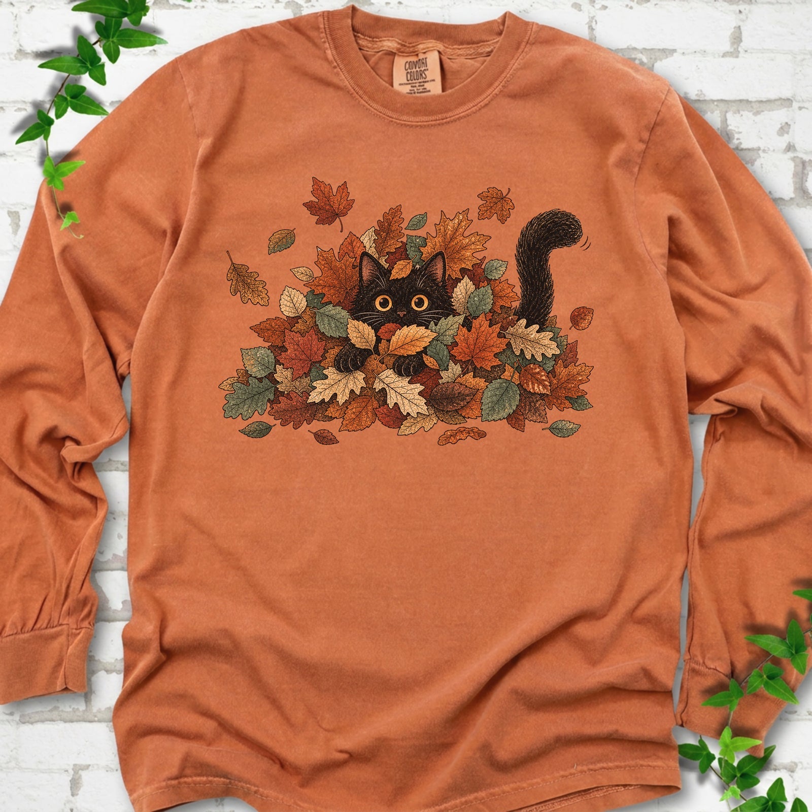 Yam Comfort Colors® long sleeve T-shirt flat lay featuring a black cat peeking from a pile of orange and green autumn leaves.