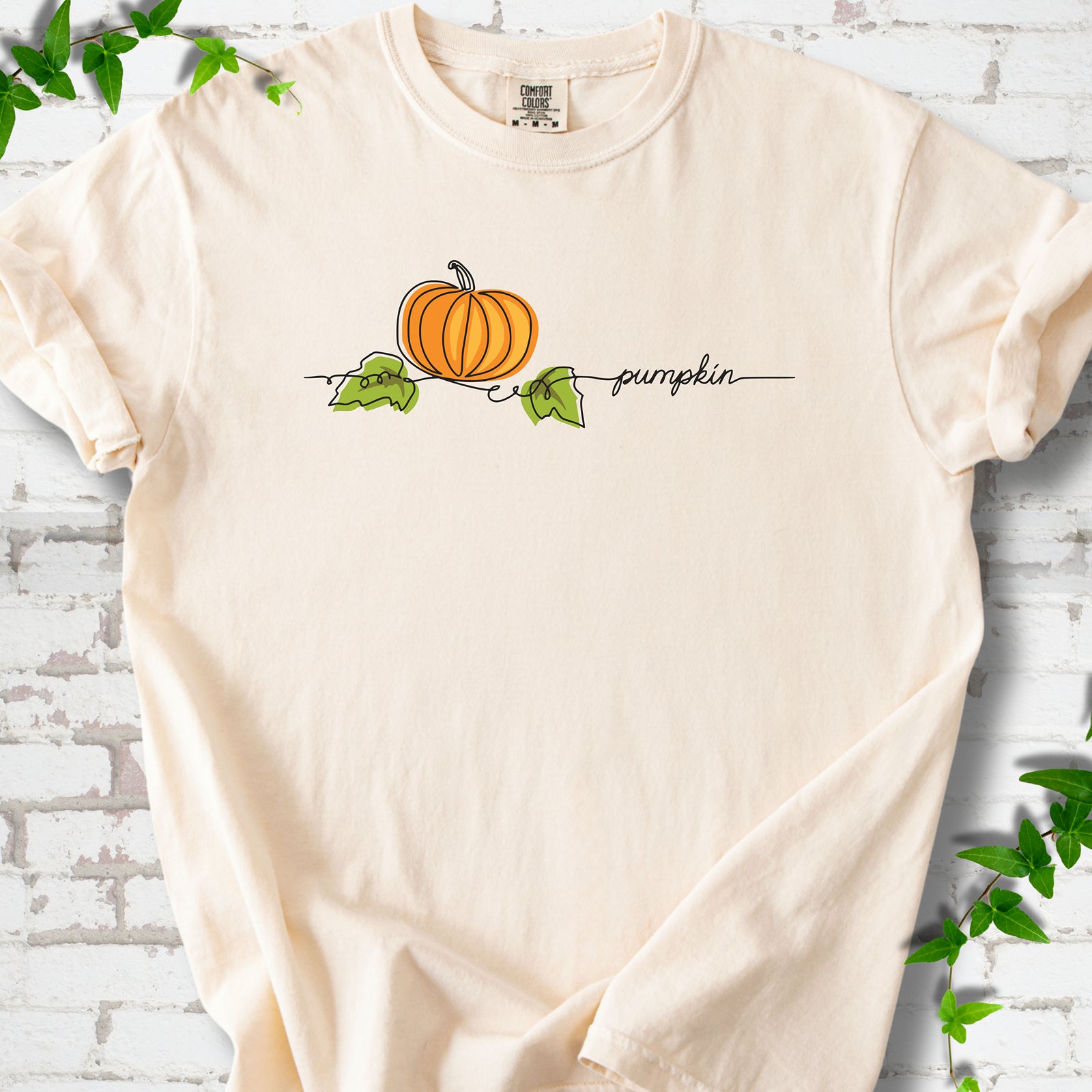Ivory Comfort Colors® T-shirt flat lay featuring a minimalist orange pumpkin, green leaves and delicate black script.