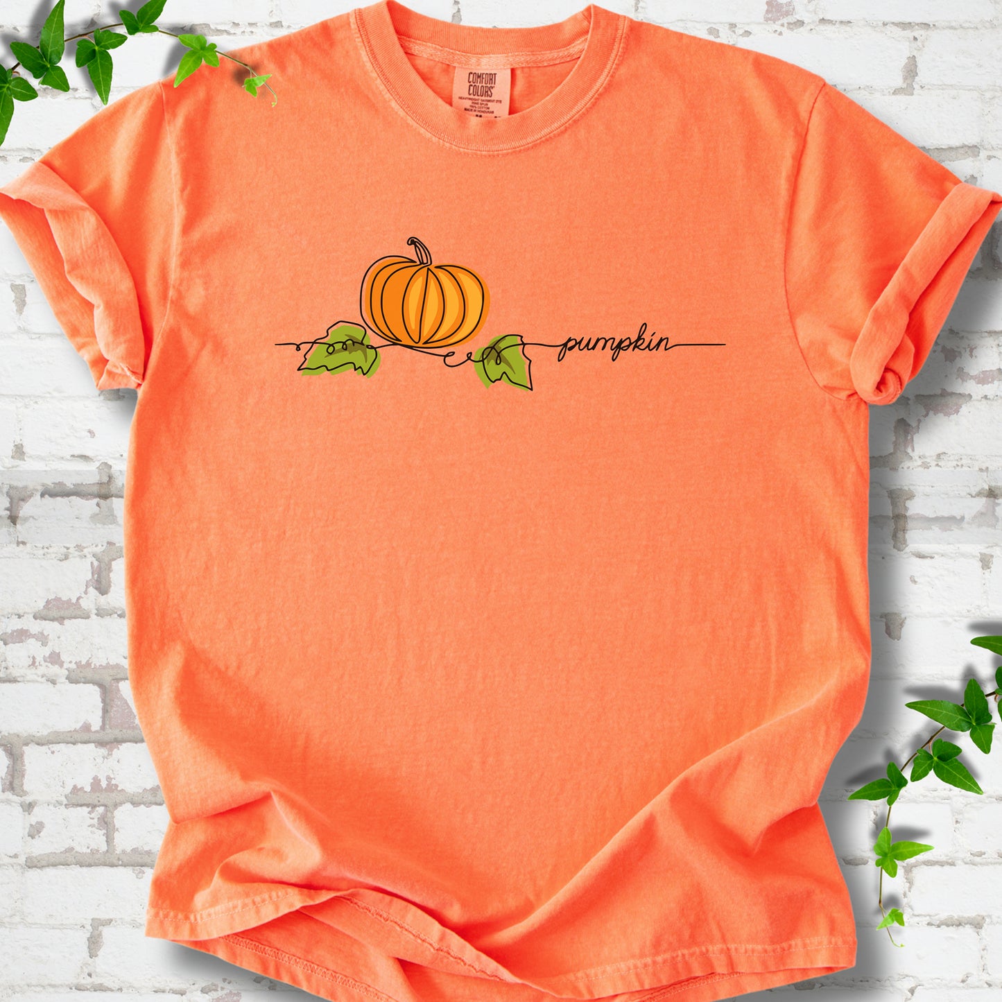 Melon Comfort Colors® T-shirt flat lay featuring a minimalist orange pumpkin, green leaves and delicate black script.