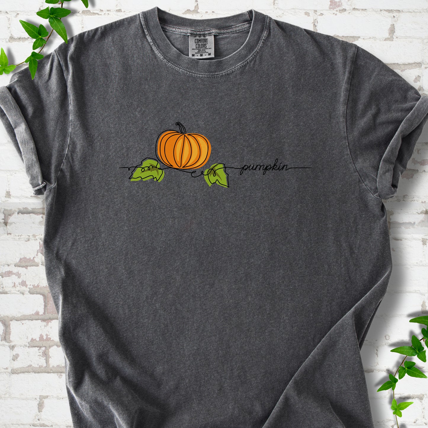 Pepper Comfort Colors® T-shirt flat lay featuring a minimalist orange pumpkin, green leaves and delicate black script.