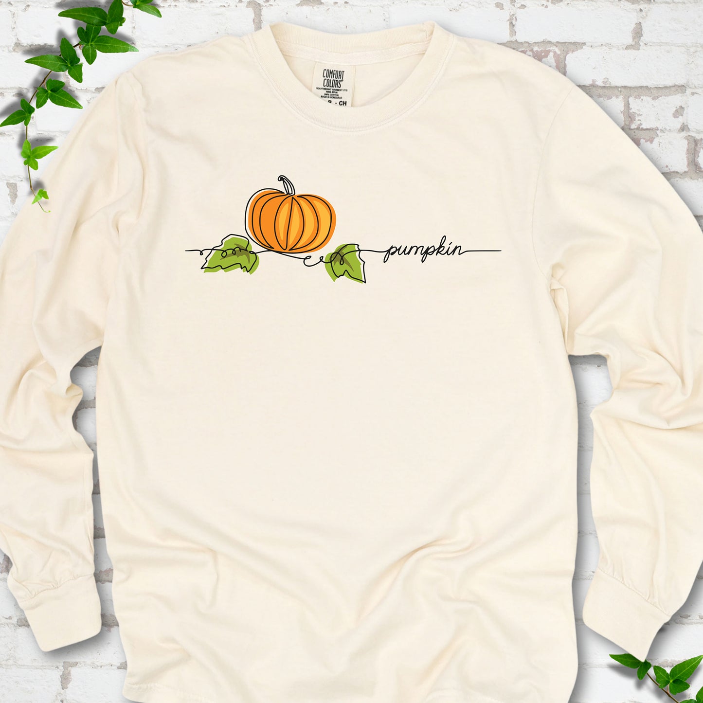 Ivory Comfort Colors® long sleeve T-shirt flat lay featuring a minimalist orange pumpkin, green leaves and delicate black script.