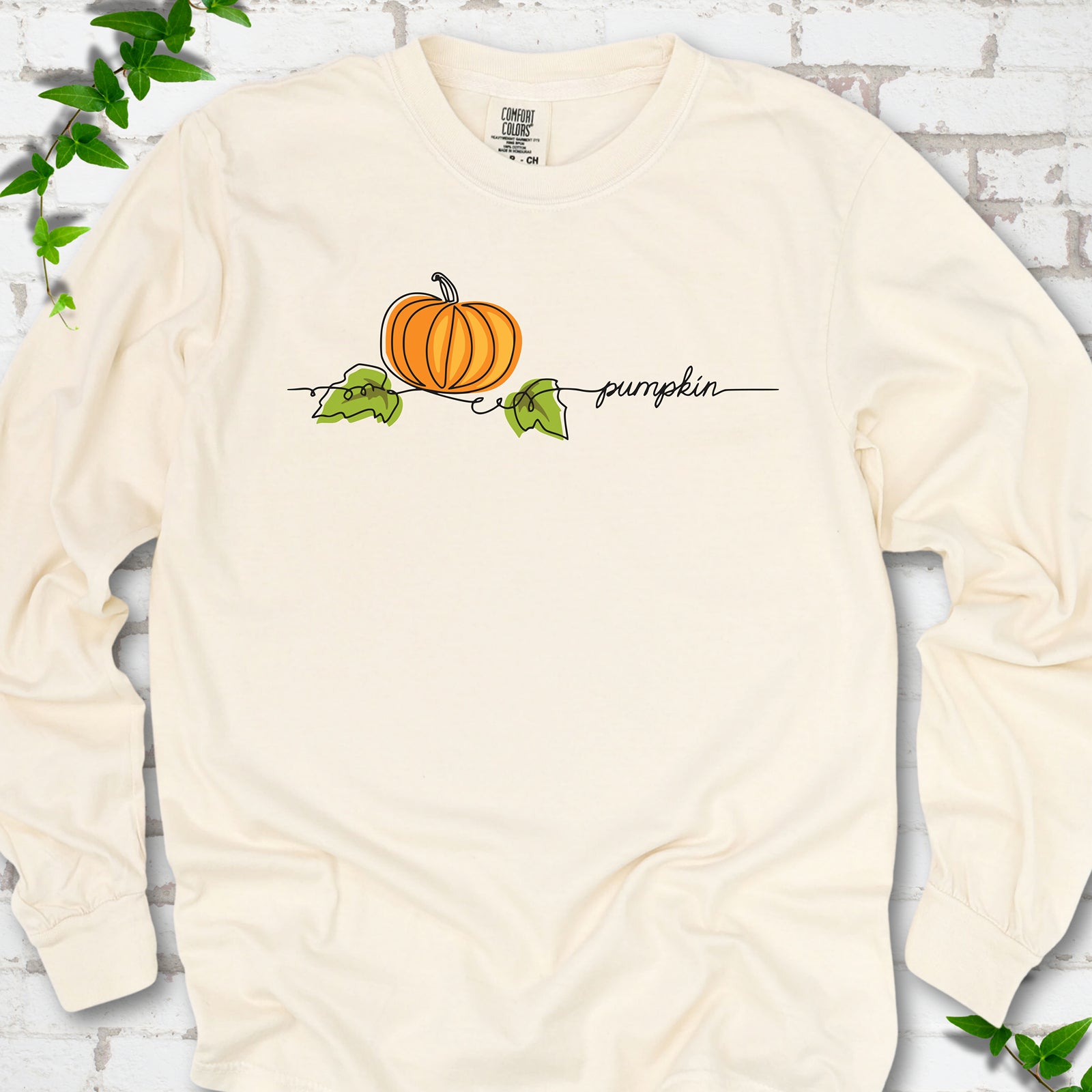 Ivory Comfort Colors® long sleeve T-shirt flat lay featuring a minimalist orange pumpkin, green leaves and delicate black script.